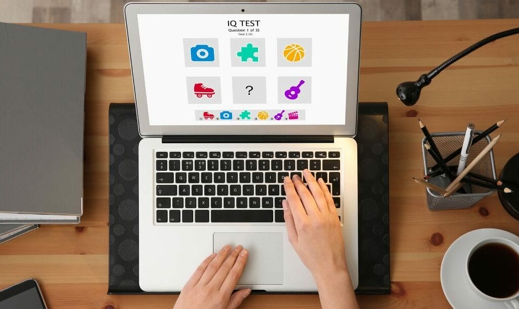 Header image, top view of a woman’s hands typing on a laptop at a desk doing an IQ test