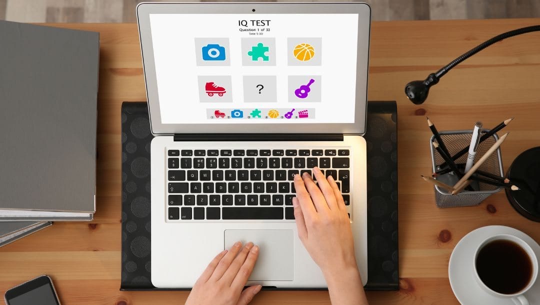 Header image, top view of a woman’s hands typing on a laptop at a desk doing an IQ test