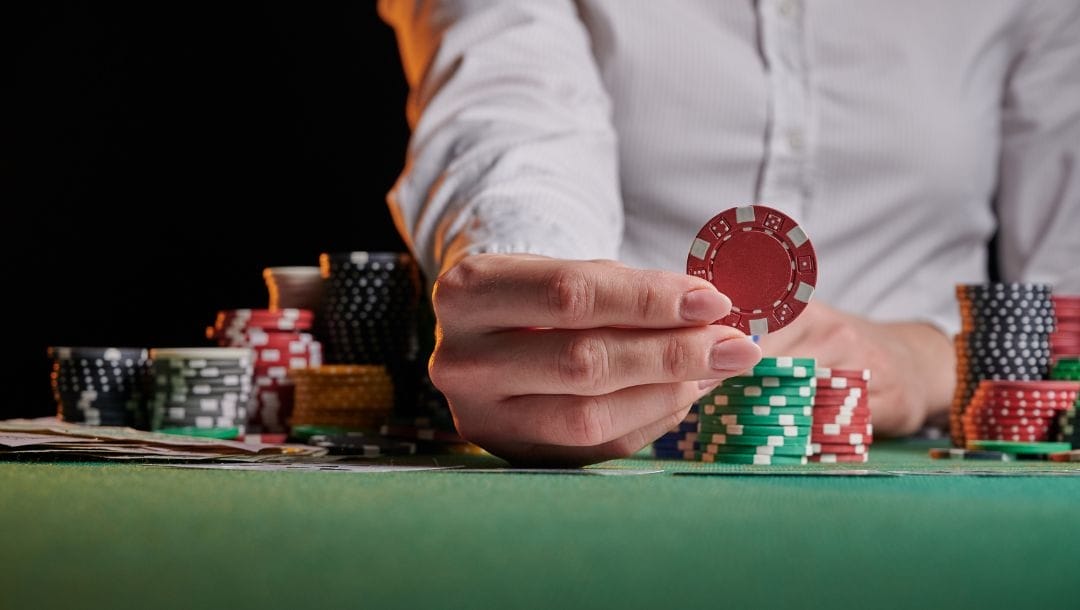 close up of a woman holding up a red poker chip on a green felt poker table stacked with playing cards and poker chips
