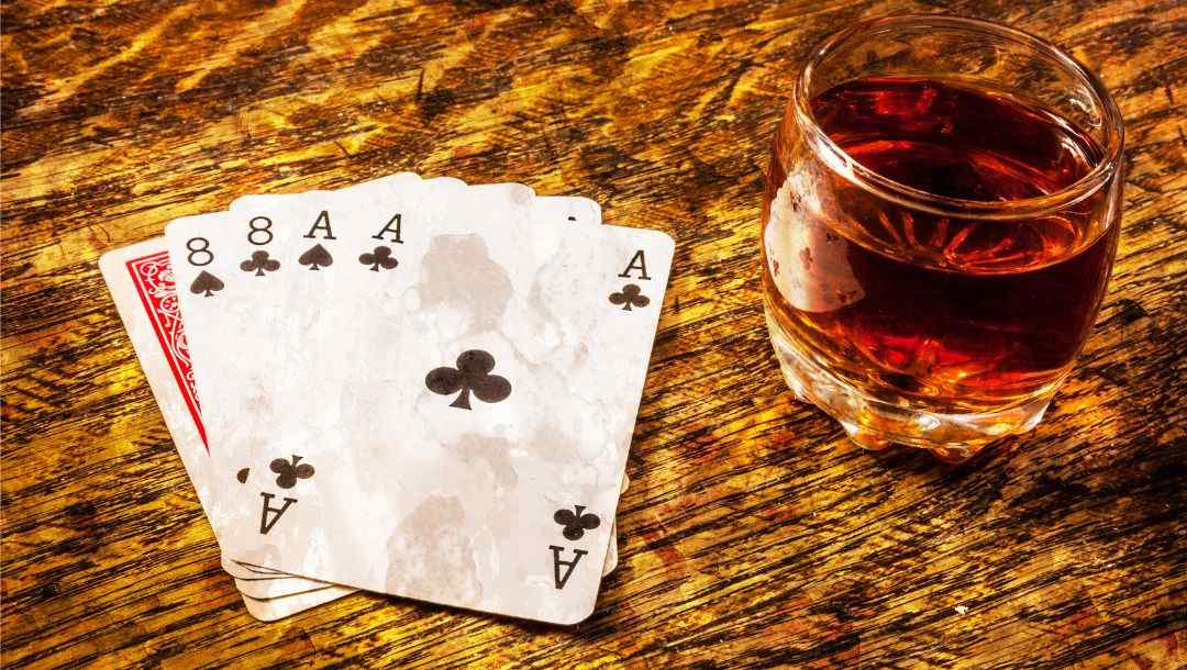 Dead Man’s Hand, two aces and two eights, on a wooden surface next to a drink in a glass