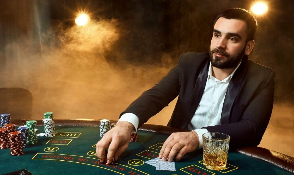 A man in a suit sitting at a green felt poker table playing poker with a drink on the table to his left and stacks of poker chips to his right, smoky background