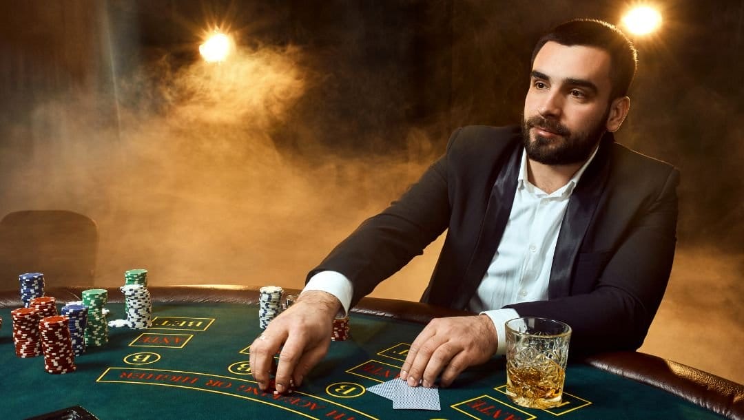 A man in a suit sitting at a green felt poker table playing poker with a drink on the table to his left and stacks of poker chips to his right, smoky background