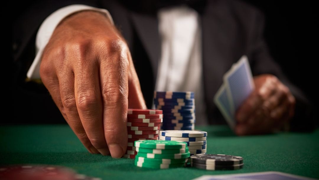 a man’s hand pushing a stack of red poker chips away from himself towards other stacks of poker chips on a green felt poker table while holding his hole cards in his other hand