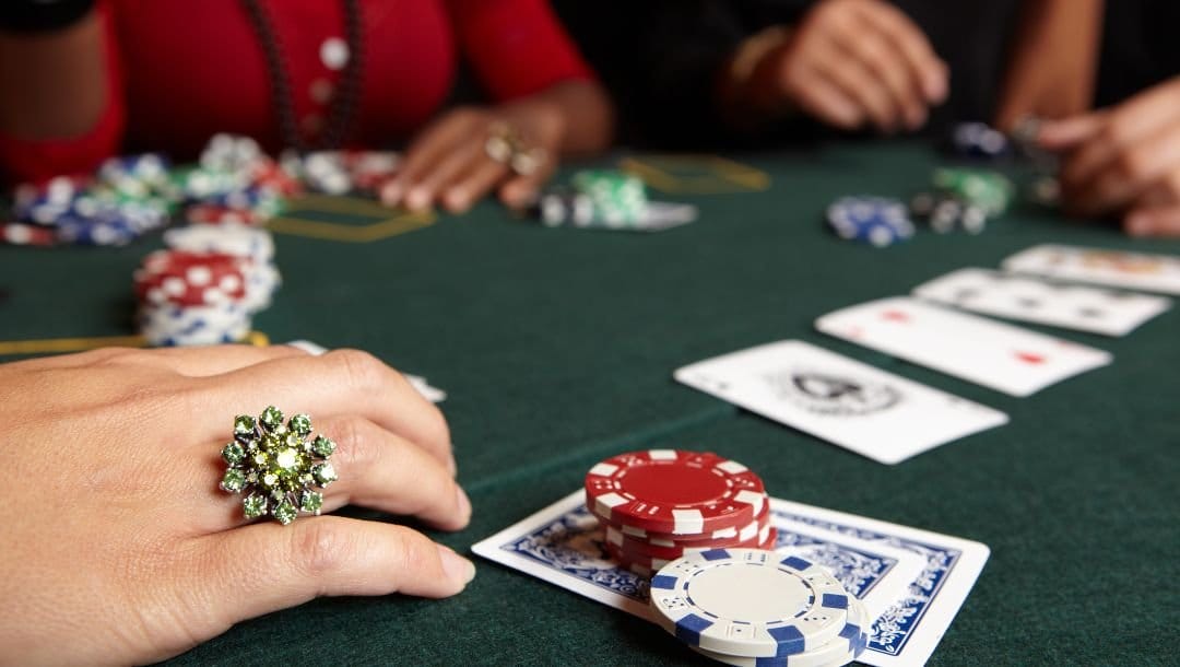 Close up of a woman’s hand wearing a decorative jewel ring resting on a green felt poker surface next to her two hole cards that have poker chips on them, in the background are other players playing poker at the same table