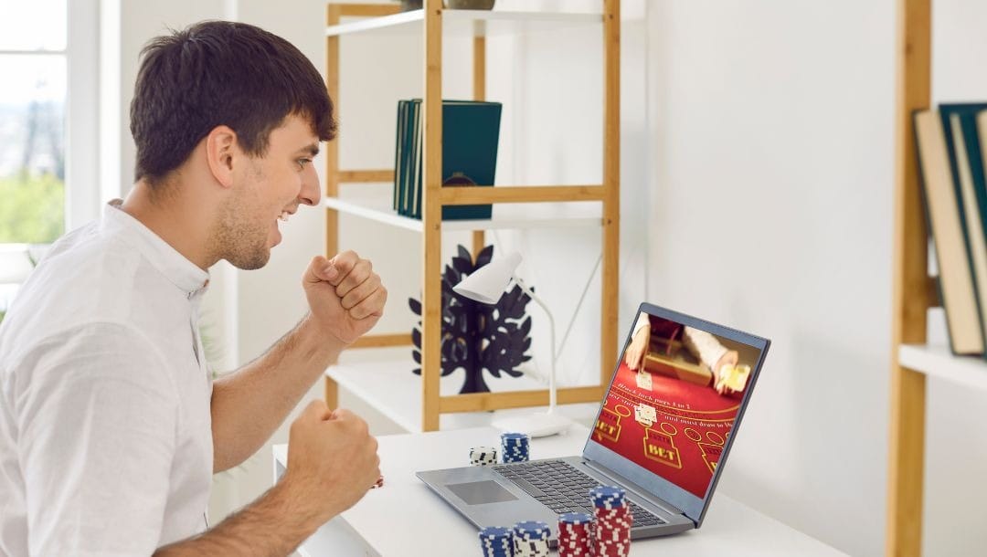 a man cheering as he sits in front of a laptop displaying online poker with stacks of poker chips stacked next to the laptop on a white desk