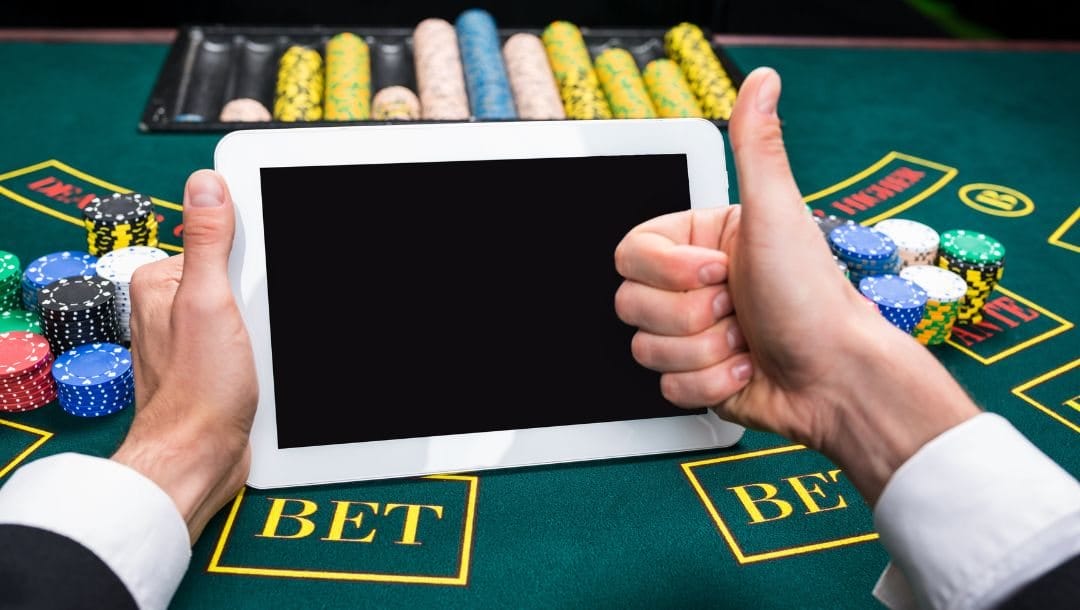 a man holding a digital tablet above a green felt poker table with poker chips stacked on it, his other hand is giving a thumbs up
