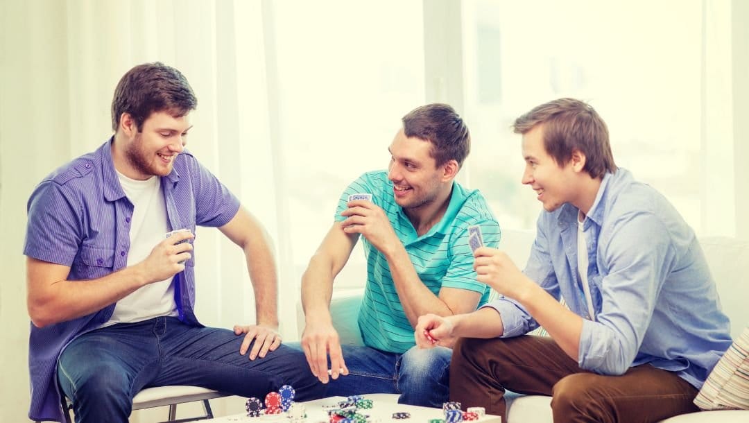 three men are playing poker at home in casual clothing, there are stacks of poker chips on the table and the two men are looking at the one whose turn it is