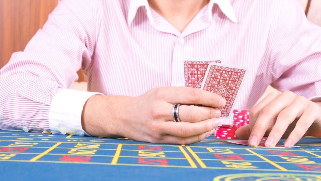 a man wearing a collared striped shirt and a silver and black ring on his finger is holding a pair of playing cards above a poker table with red poker chips on it