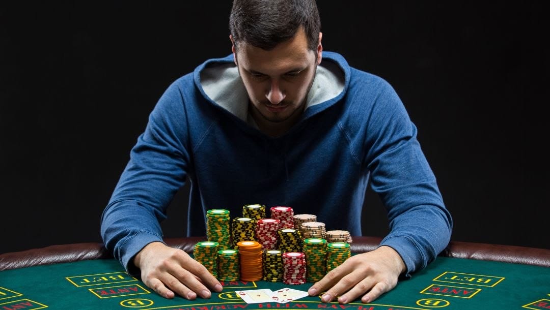 a man in a blue hooded sweater is sitting at a poker table with stacks of poker chips in front of him as he stares at his two hole cards on the table, a pair of aces