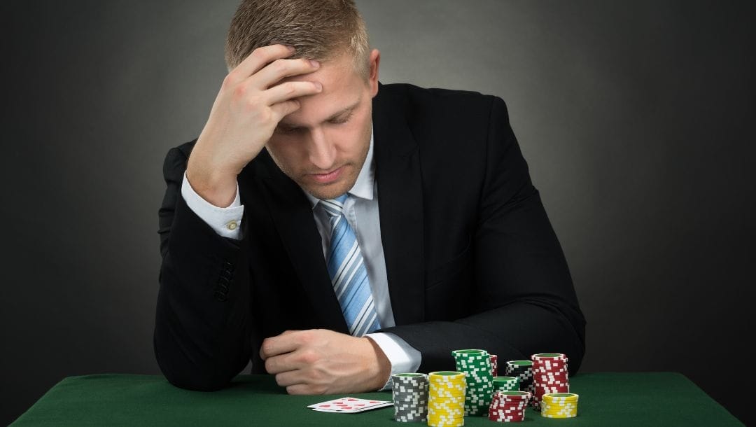 a man in a suit and tie is sitting at a small poker table with his head down and his hand on his forehead, in front of him are two playing cards and stacks of poker chips