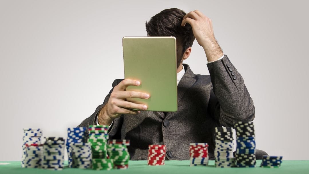 a man wearing a suit is holding up a notebook in front of his face and scratching his head with his other hand, on the green felt poker table in front of him are stacks of poker chips