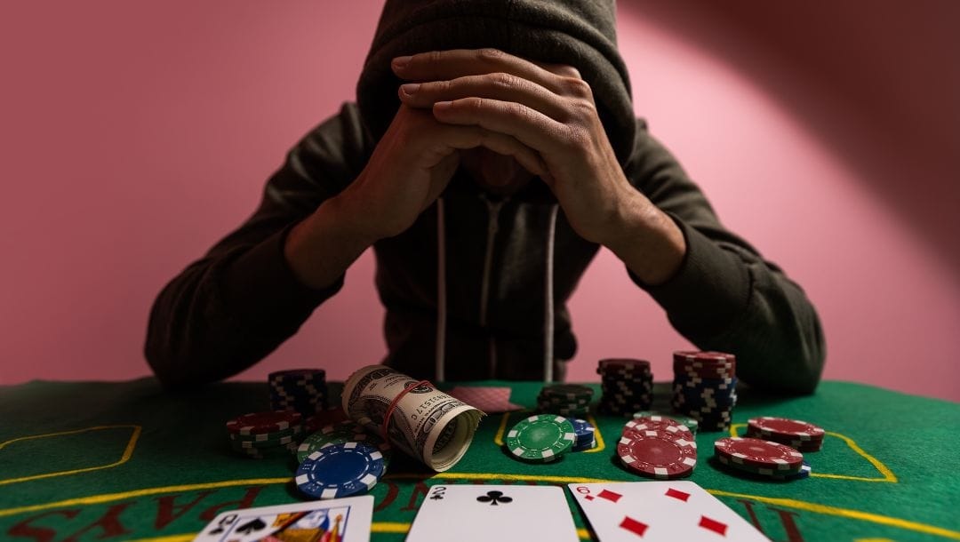 a man in a hooded sweatshirt is sitting at a green felt poker table with his head down and both his hands on his forehead, on the table in front of him are poker chips, playing cards and some money