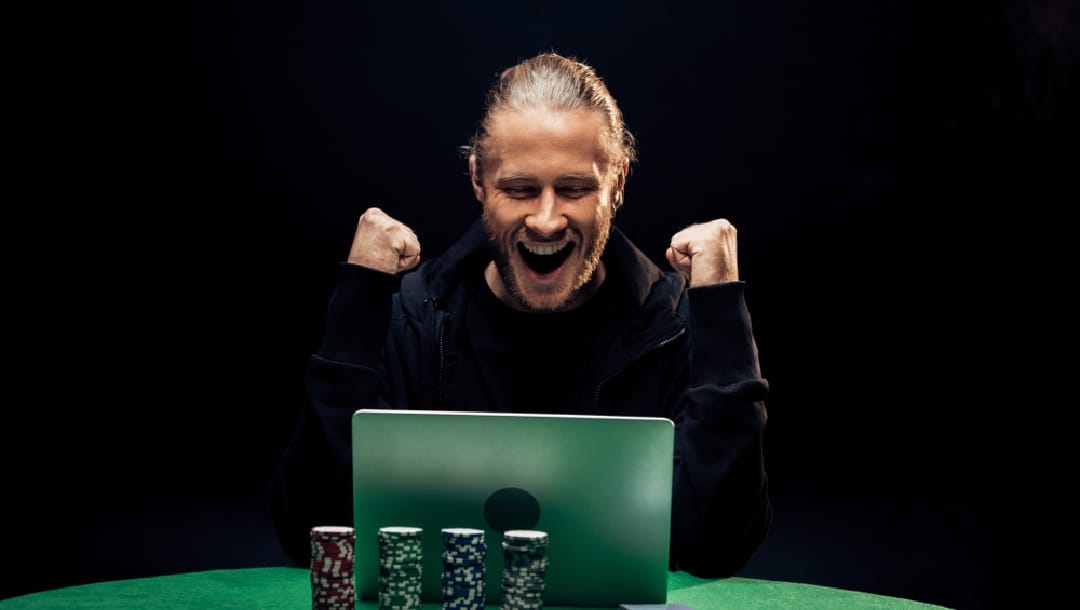 A man celebrates winning on his laptop while sat at a poker table with poker chips behind him