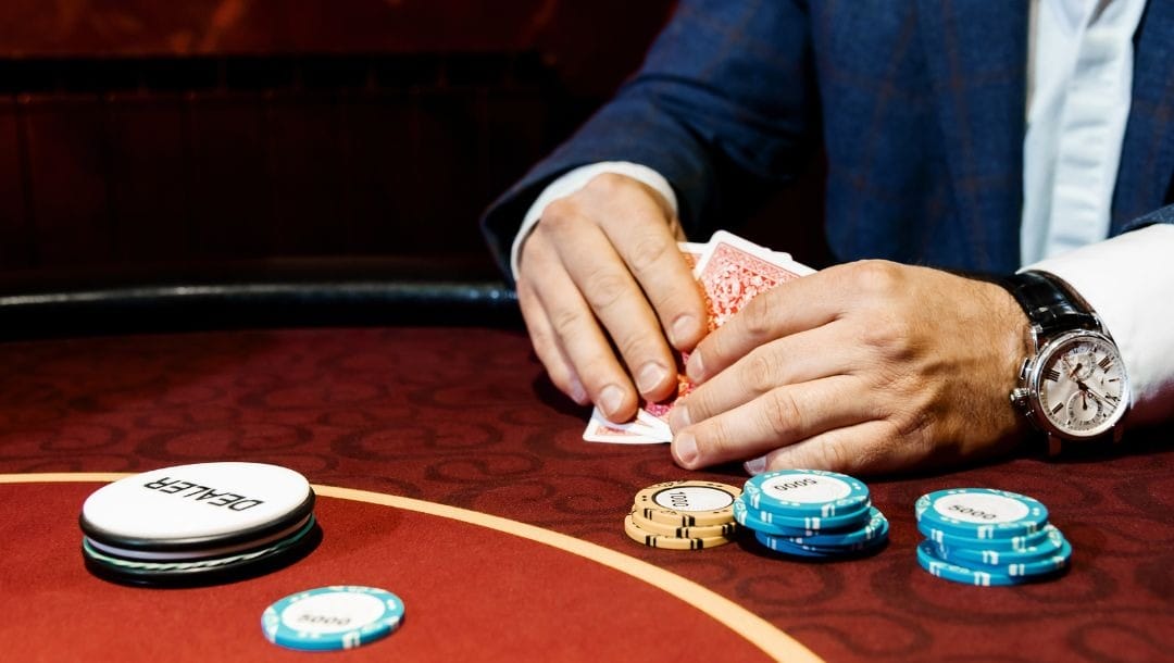 a close up of a man checking his hole cards during a game of poker at a casino, in front of him on the red felt poker table are poker chips and a dealer token