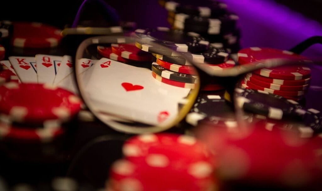 a pair of glasses magnifying a royal flush of hearts hand of playing cards on a table that has stacks of red and black poker chips on it