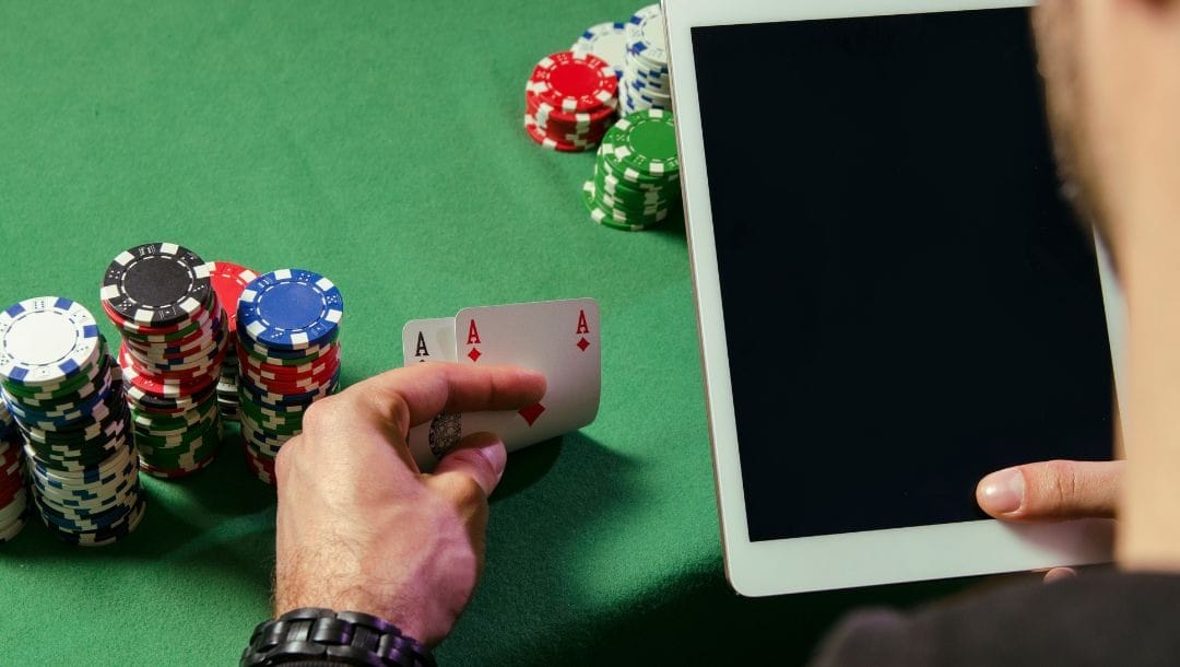 an over-the-shoulder view of a man holding a digital tablet in his right hand and a pocket pair of ace playing cards in his left hand while sitting at a green felt poker table with poker chips stacked on it