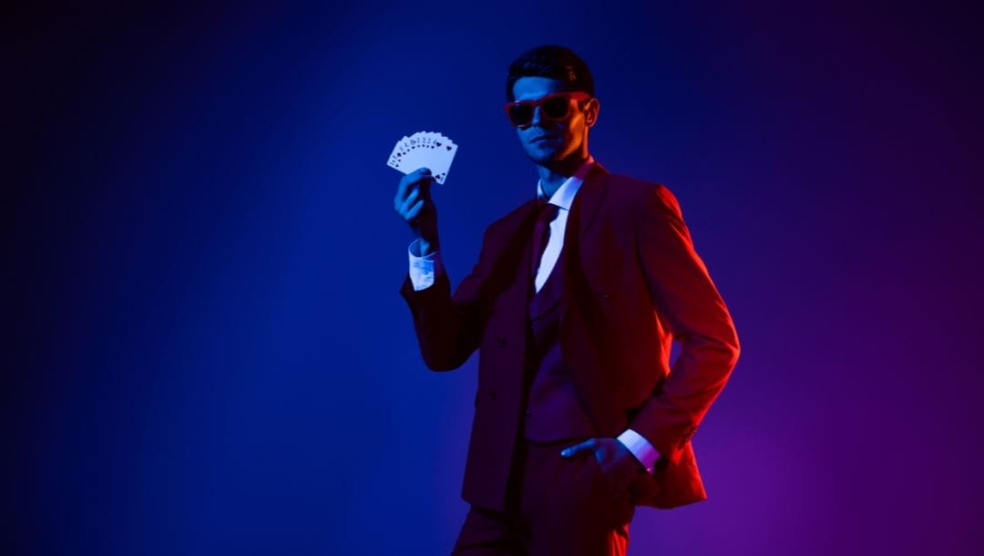 a man in a suit and sunglasses is holding up a hand of playing cards fanned out with a dark blue background