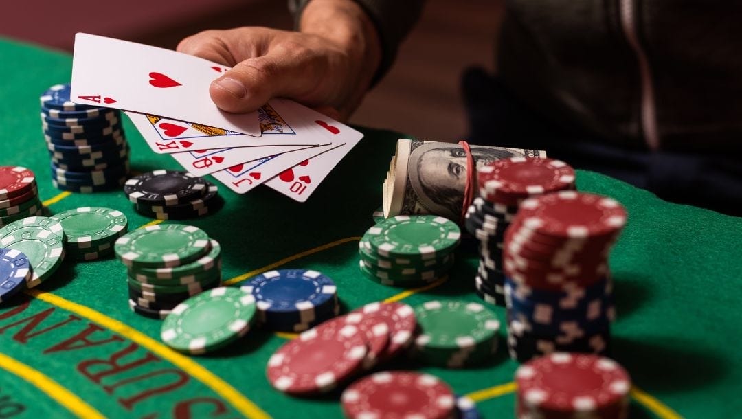 close up of a man holding a royal flush of hearts above a green felt poker table with money and stacks of poker chips on it