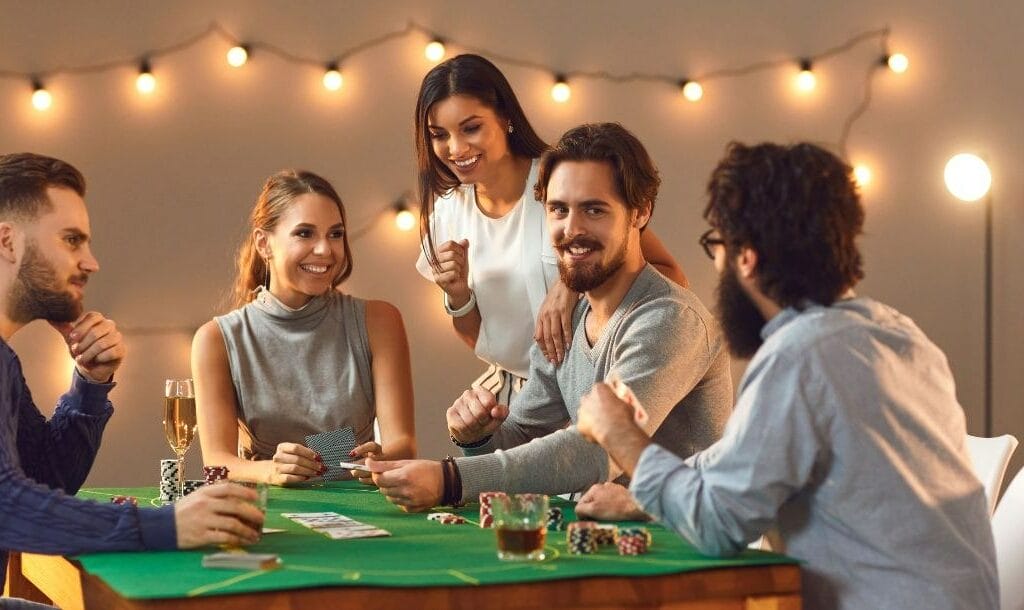 five friends are playing poker while gathered around a dining table that has been turned into a poker table with a green felt table cover, playing cards and poker chips on it, in the background are hanging lights on the wall