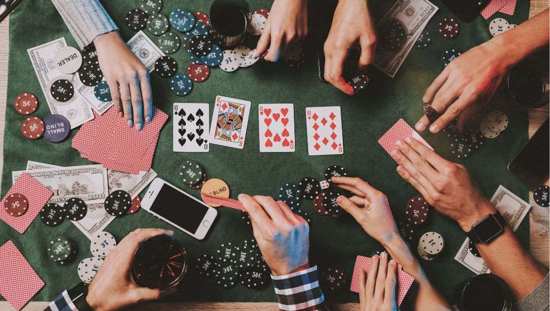 aerial shot of an at-home poker table with people gathered around it playing poker, on the table there are poker chips, playing cards, dealer and blind tokens, drinks, cellphones and money