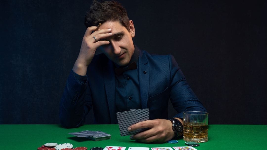 a man in a suit with a distressed expression is holding a cigar and looking at his two hole cards at a poker table that has poker chips, playing cards and a whiskey glass on it