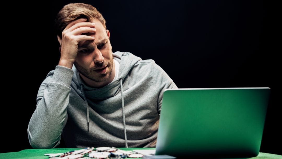 a man with an angry and confused facial expression is sitting at a poker table looking at an open laptop with a pile of poker chips next to it