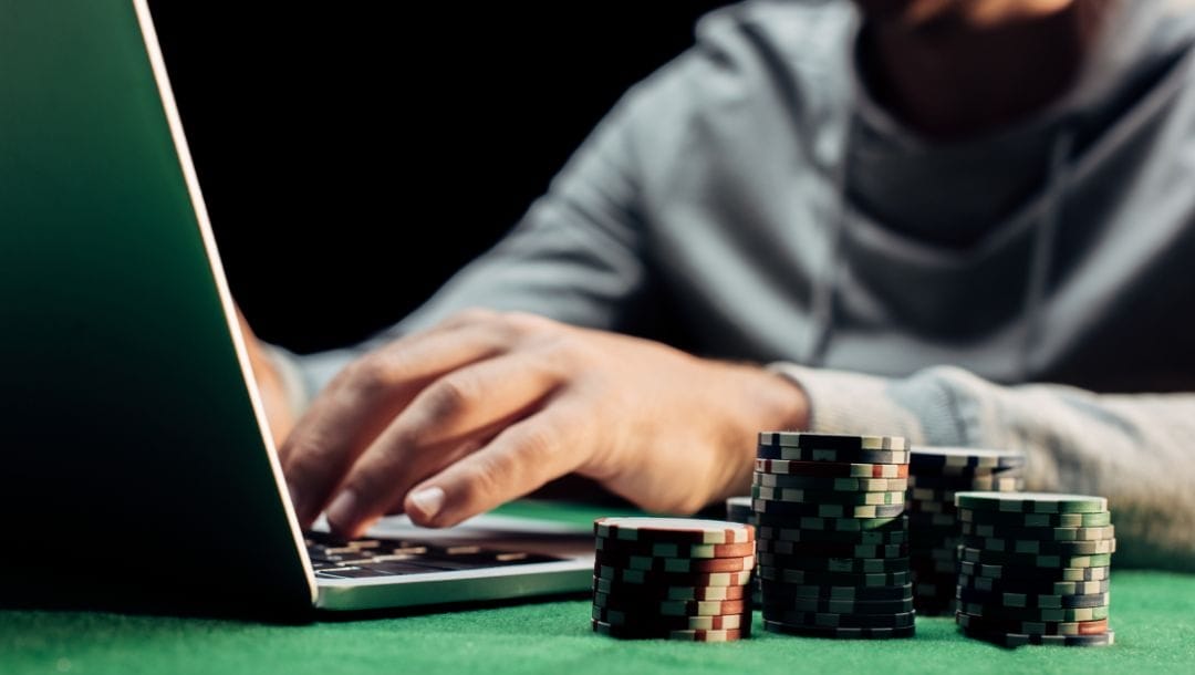 A person typing on a laptop on a green felt poker surface with poker chips stacked next to it