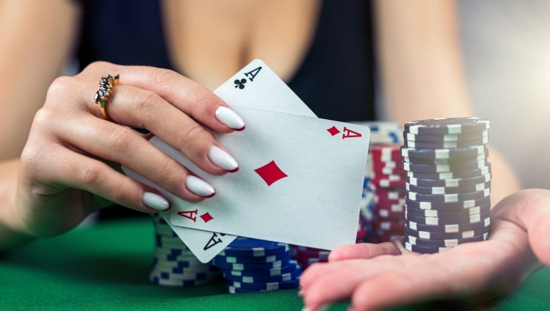 a woman with perfectly manicured hands wearing a ring is holding up a pair of ace playing cards, a club and a diamond, in her right hand and a stack of blue poker chips in the palm of her left hand above a green felt poker table with other stacks of poker chips on it