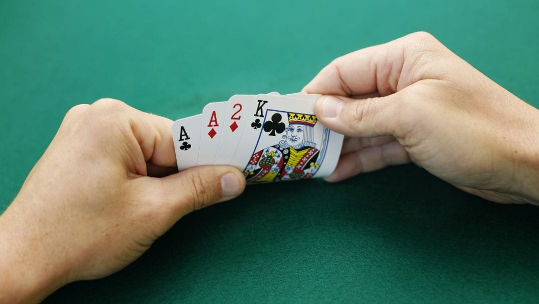 a person is holding four playing cards, an ace and king of clubs and an ace and two of diamonds, above a green felt poker surface