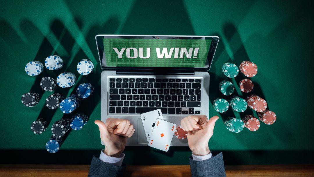 a top view shot of a man winning an online poker game, he is sitting at a green felt poker table with his thumbs up, an open laptop in front of him that says “YOU WIN”, poker chips stacked on either side of the laptop and two ace playing cards on the laptop keyboard
