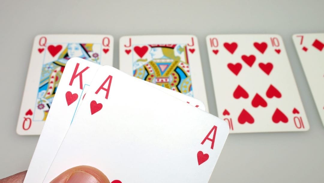 A person’s hand holding up two playing cards, an ace and a king of hearts, with four playing cards on the surface behind, a queen, a jack, a ten of hearts, and a seven of diamonds, indicating a royal flush altogether.