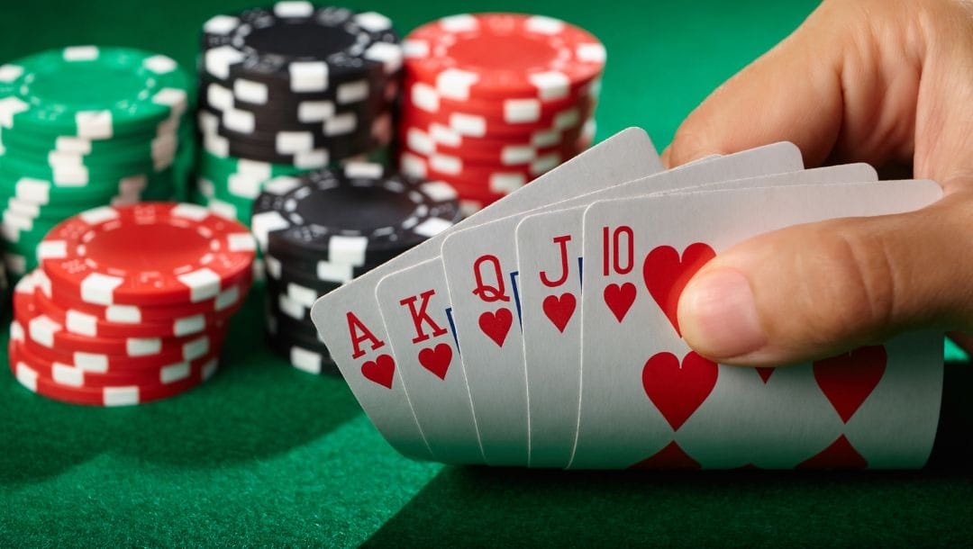 A person flipping up the edges of five playing cards, a royal flush of hearts, on a green felt poker surface with stacks of poker chips behind them.