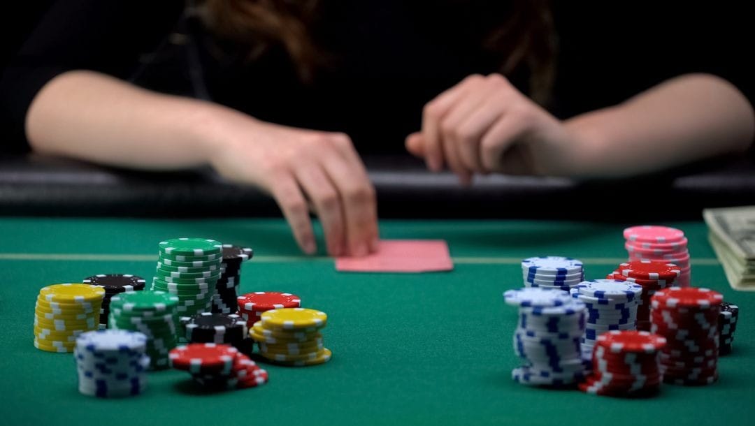 a woman sitting at a green felt poker table with poker chips stacked on it, she is about to flip her hole cards to look at them