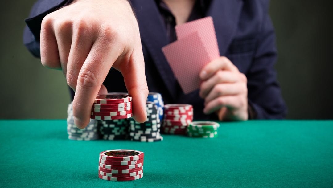 A poker player holding two cards picks up a small stack of red poker chips and is about to place them on another.