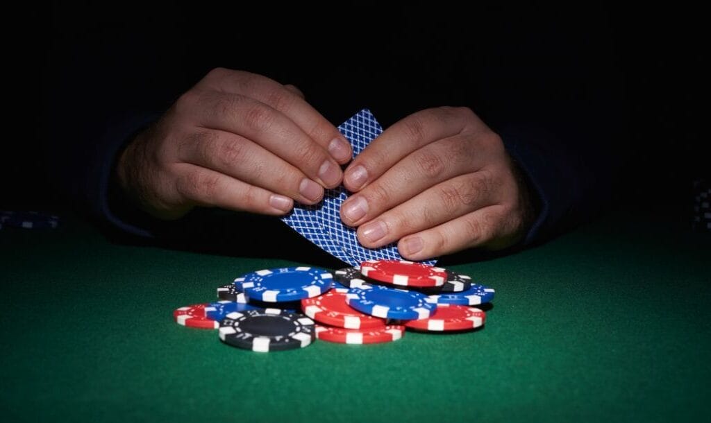 a close up of a man’s hands holding two playing cards coming out of the shadow towards the poker chips on the green felt poker surface