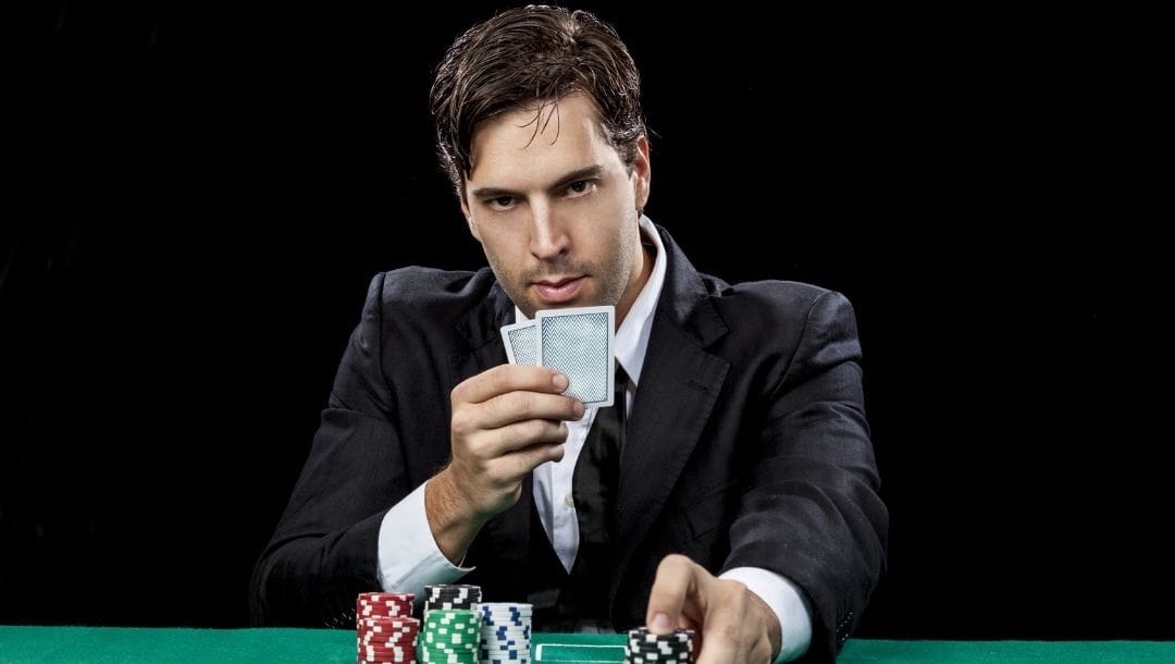 a man holds his two hole cards up to his face secretively and pushes a stack of black poker chips forward on a green felt poker surface