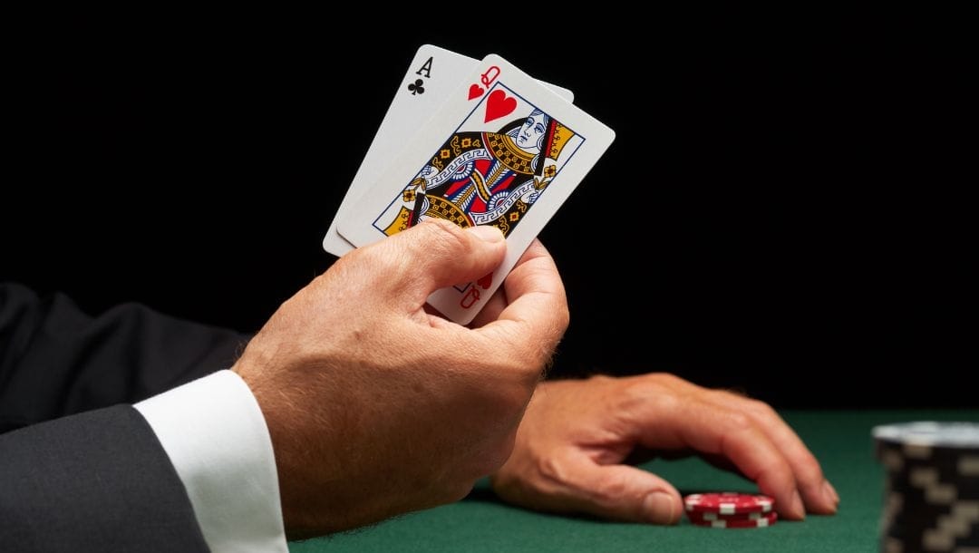 a man showing his hole cards, an ace of clubs and a queen of hearts, with his right hand and picking up poker chips with his left hand off a green felt poker surface