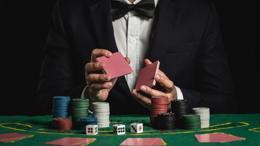 a dealer wearing a suit is shuffling a pack of playing cards with stacks of poker chips and dice in front of him on the blackjack table