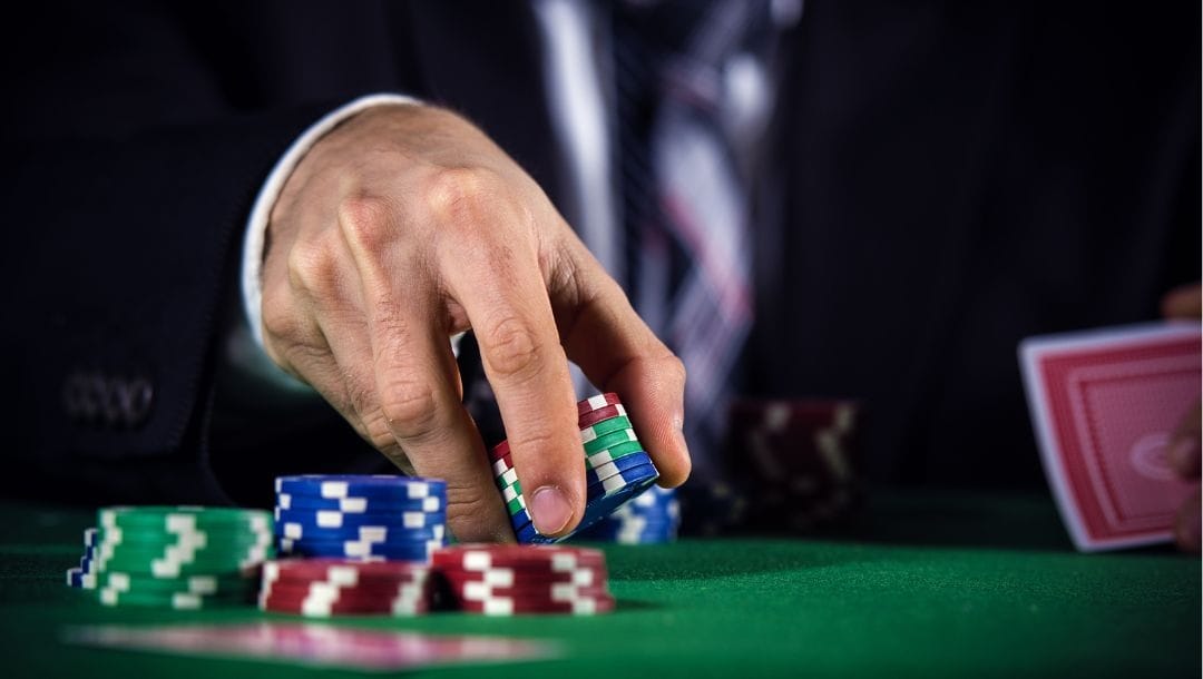 a man picking up one of several stacks of poker chips on a green felt poker table while holding playing cards in his other hand