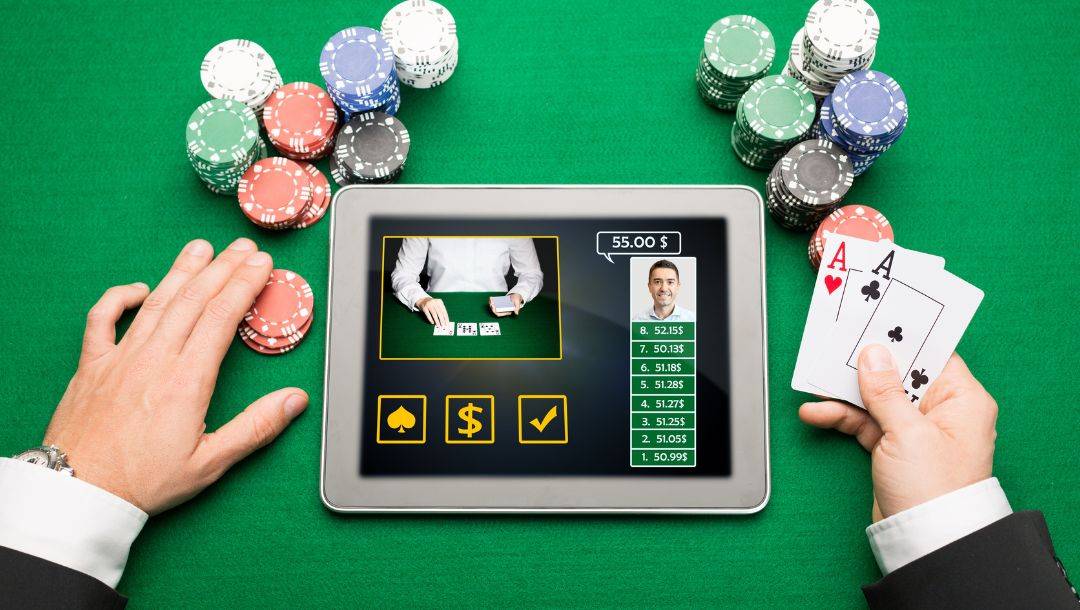 a top view close up of a man playing online poker on a digital tablet with pocket aces in his right hand and stacks of poker chips on the green felt poker surface