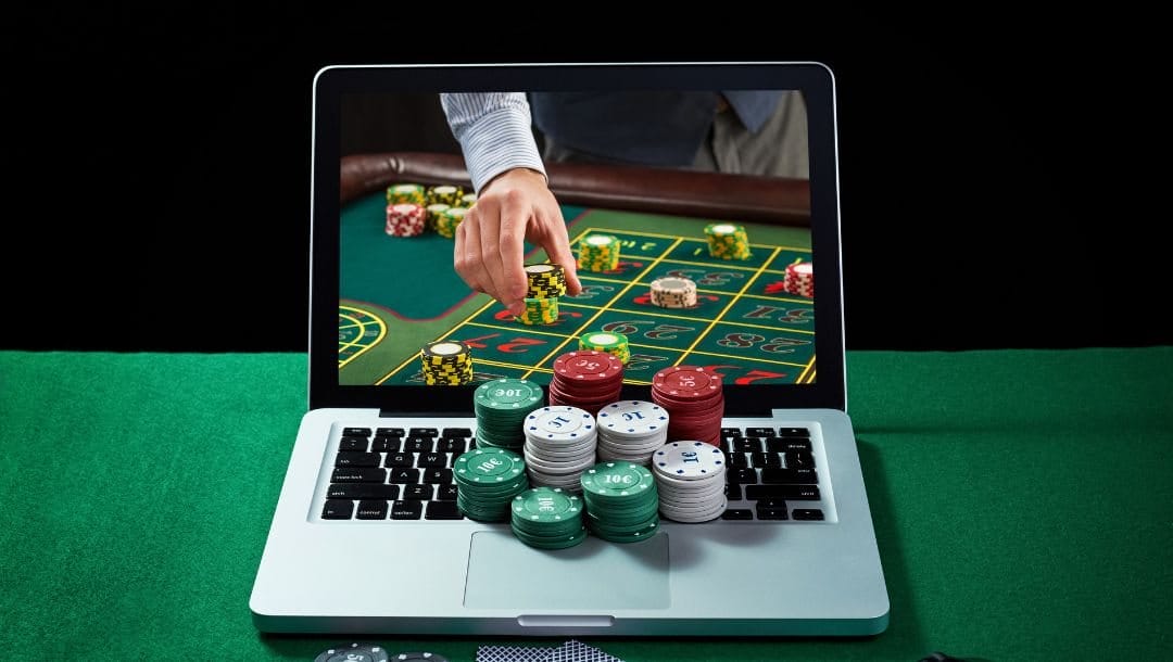 a laptop on a green felt poker surface with stacks of poker chips on its keyboard and a man betting on roulette on the screen