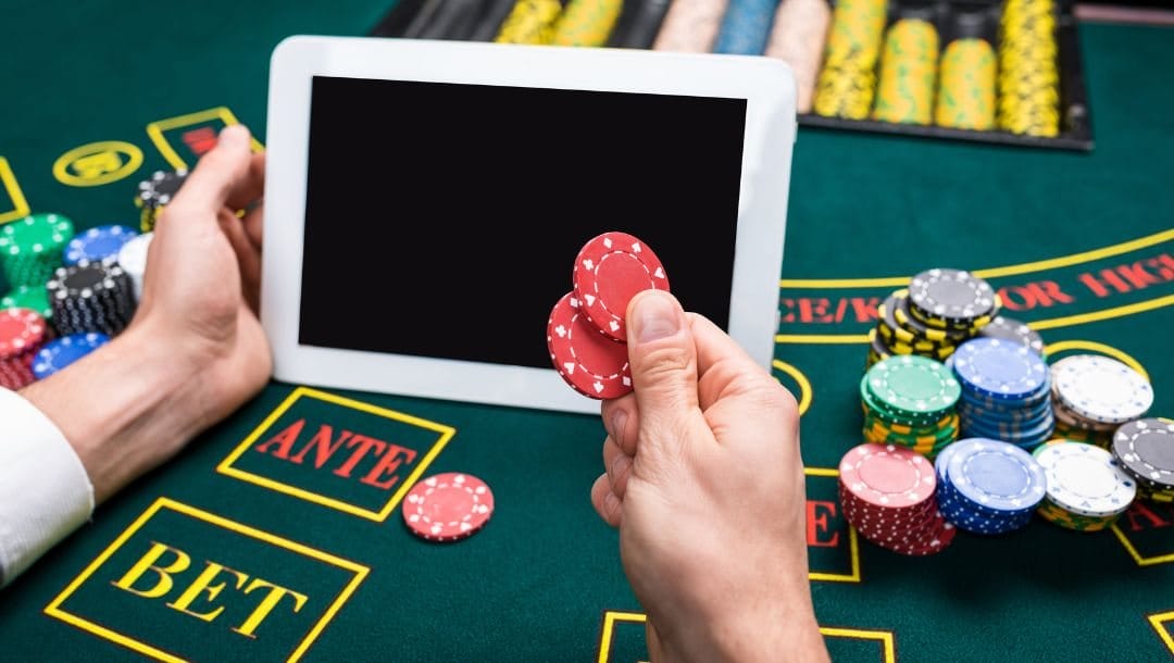 a person sitting at a blackjack table stacked with poker chips, holding two red poker chips in their right hand and a digital tablet in their left hand