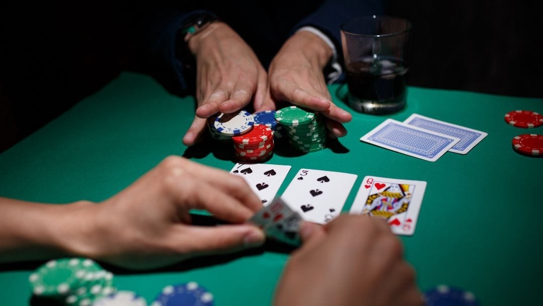 two players sitting opposite each other at a poker table, one player checks his hole cards and the player opposite him pushes all his poker chips forward towards the middle of the poker table