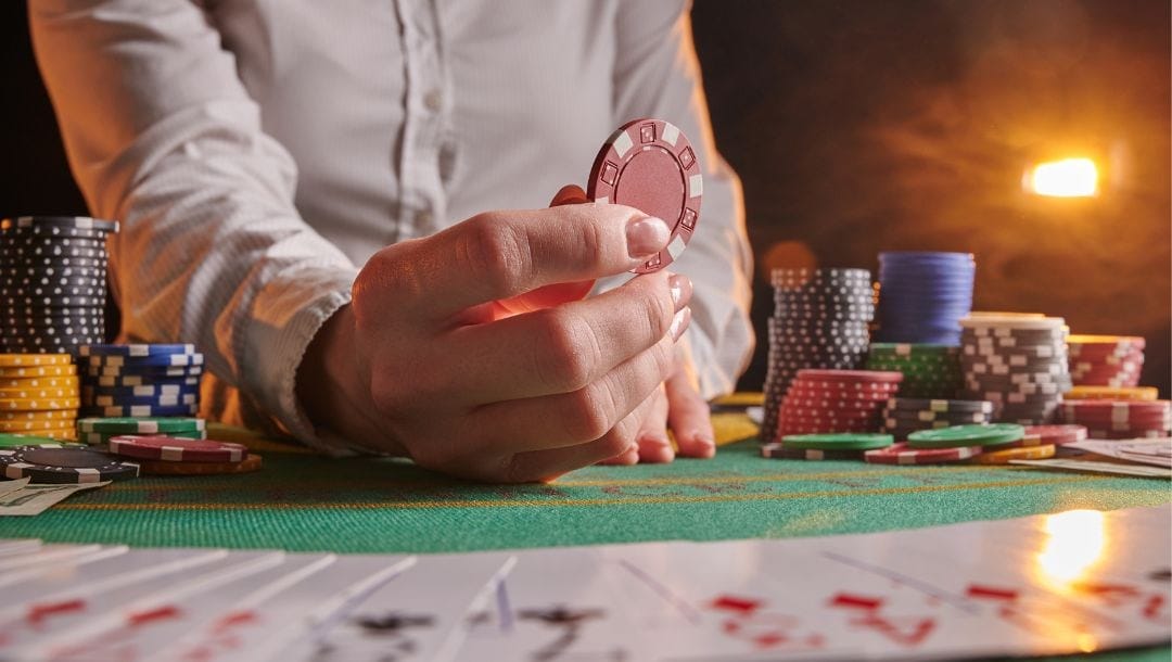 a close up of a dealer at a poker table holding up a red poker chip with stacks of poker chips on either side of them and playing cards fanned out in front of them