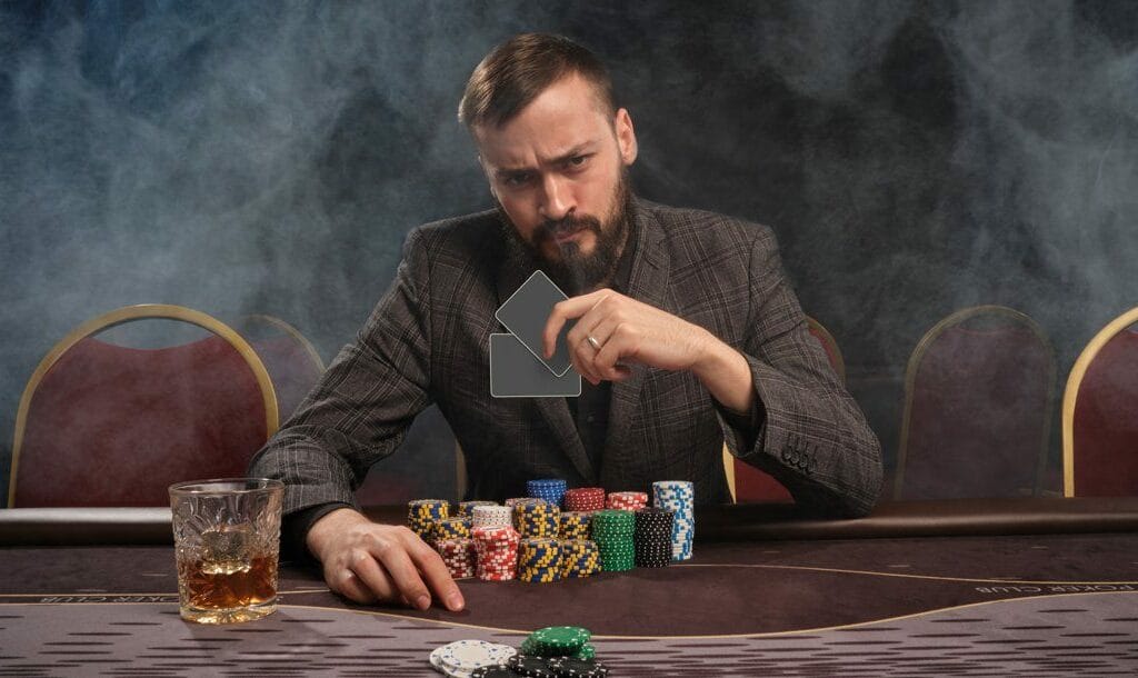 A poker player looks over at his opponent while holding his two hole cards in his hand. There are many stacks of poker chips in front of him and a drink near his right hand.