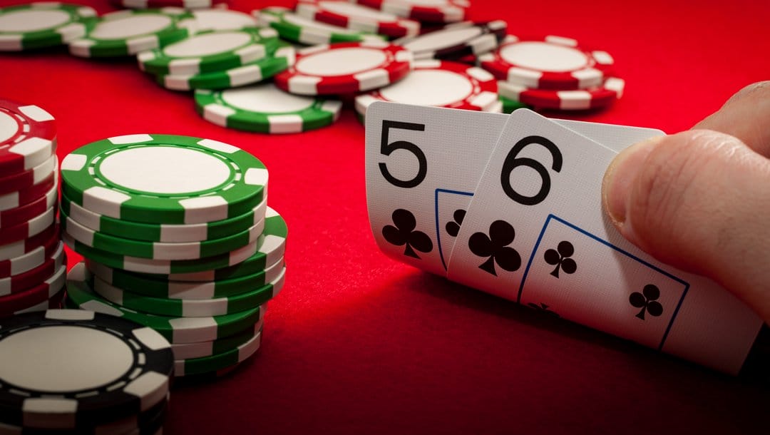 A poker player checks their hole cards. They have a five and six of clubs. There are stacks of poker chips and poker chips scattered on the table near his hand.