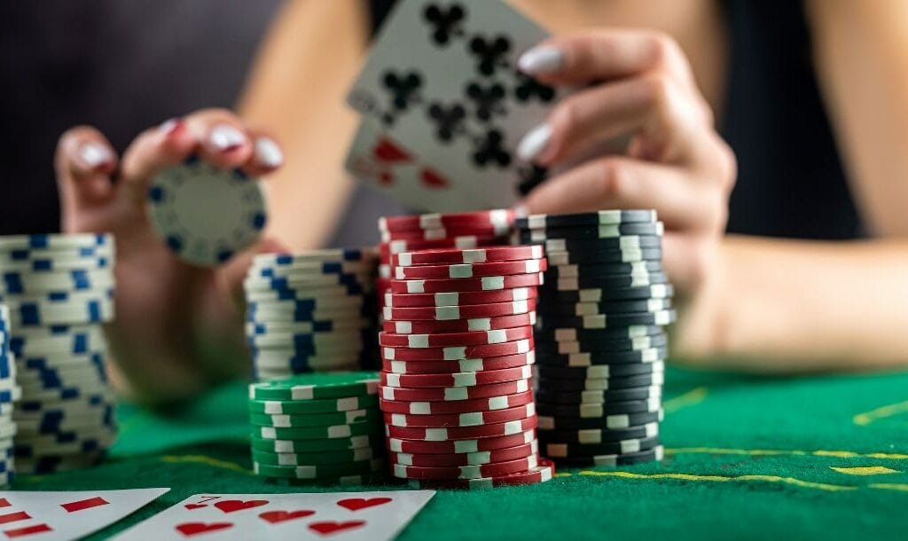 A female poker player takes a blue chip off her stack of poker chips on a green felt table.