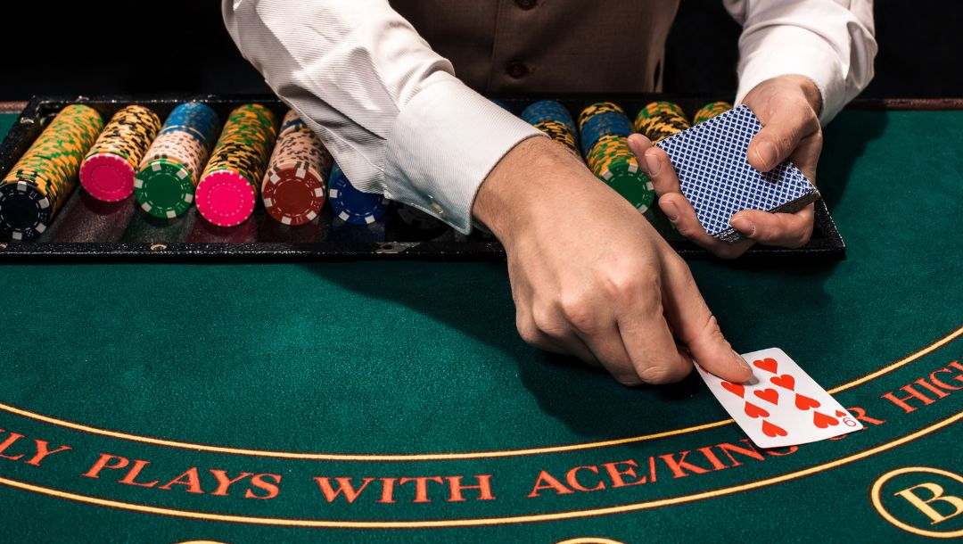 A poker dealer reaches over the chip tray to place a 9 of hearts face up on a green felt table.