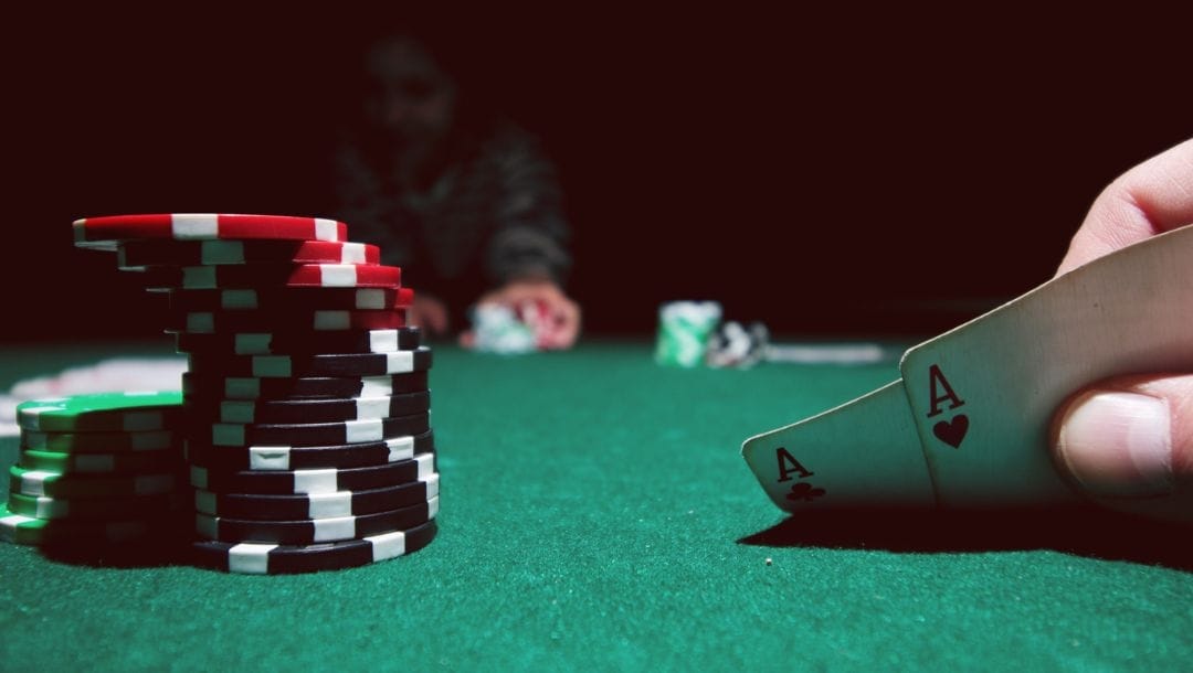 A person's hand holding two aces, surrounded by a poker table with other players engaged in a game.