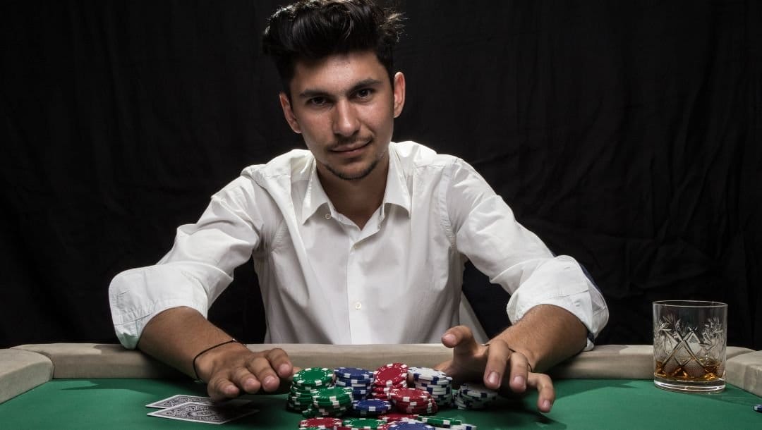 a man is sitting at a poker table that has a drink and playing cards on it and he is using both hands to push a pile of poker chips forward